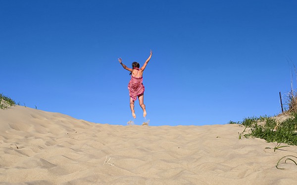 A woman in a pink dress jumps for joy against a backdrop of brilliant warm blue sky and golden sand dunes.